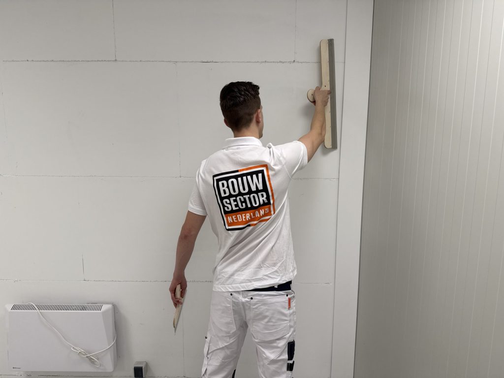 Stukadoor werkt met een lange spaan hoog op een wand, onder hem een elektrische kachel tegen de muur