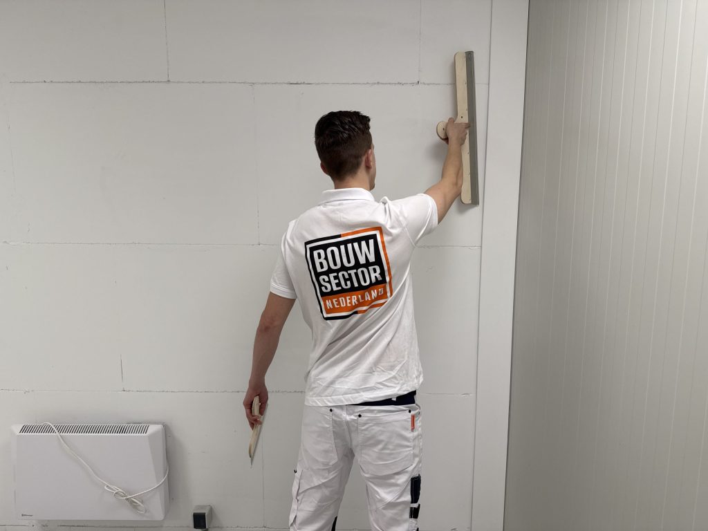 Stukadoor in wit werkshirt met het logo van Bouwsector Nederland zet een spaan boven zijn hoofd tegen de wand
