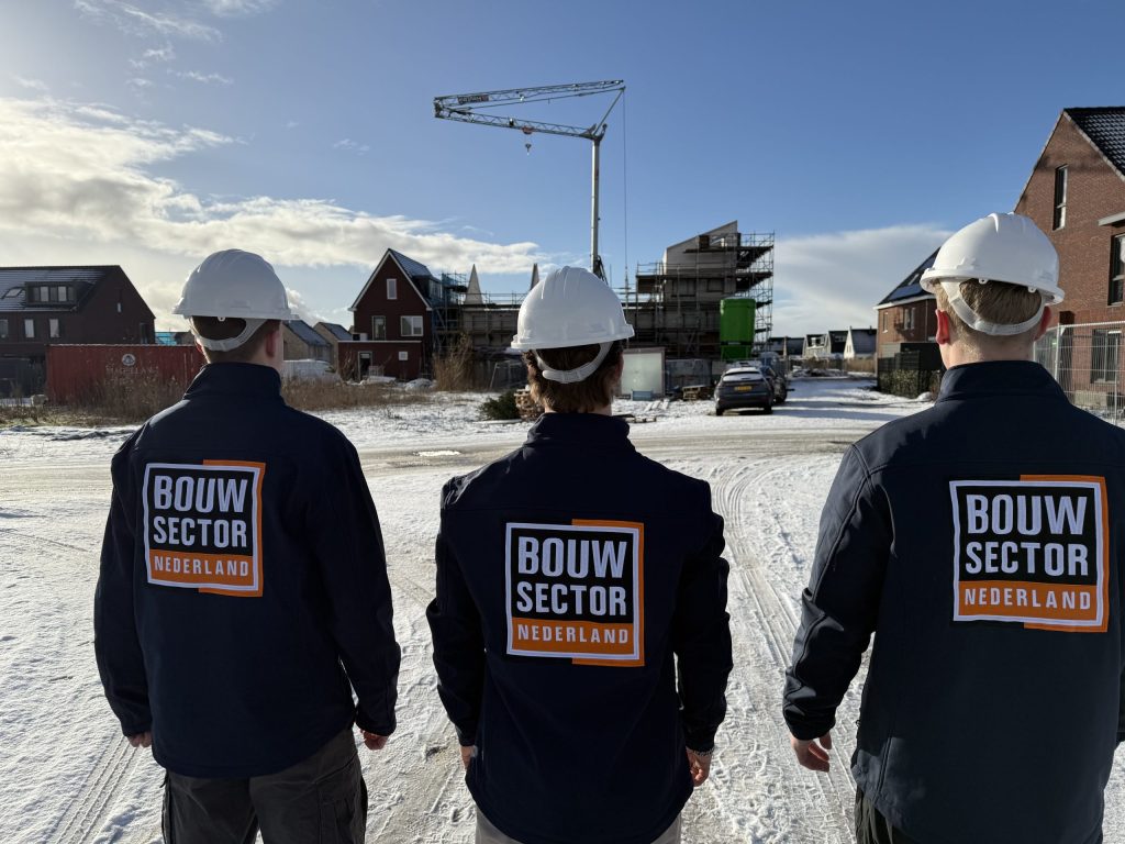 Drie vakmensen van Bouwsector Nederland met witte helm staan in de sneeuw voor een straat met huizen in aanbouw